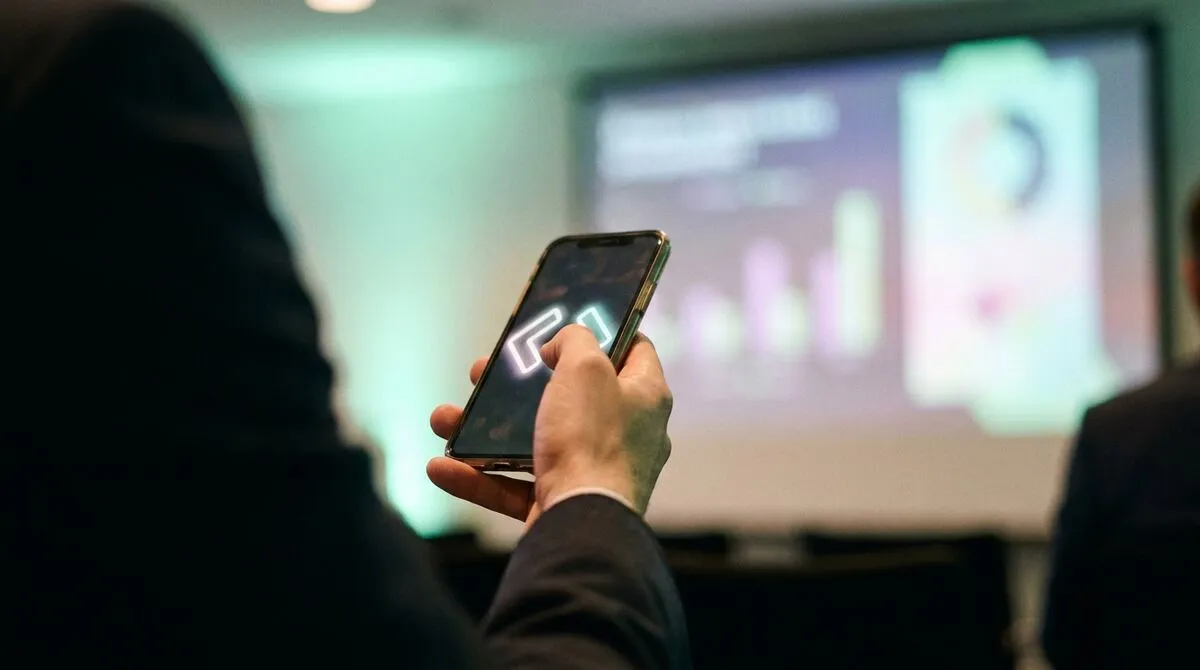 Hand holding a phone with large forward and back buttons, presentation screen glowing softly in the background