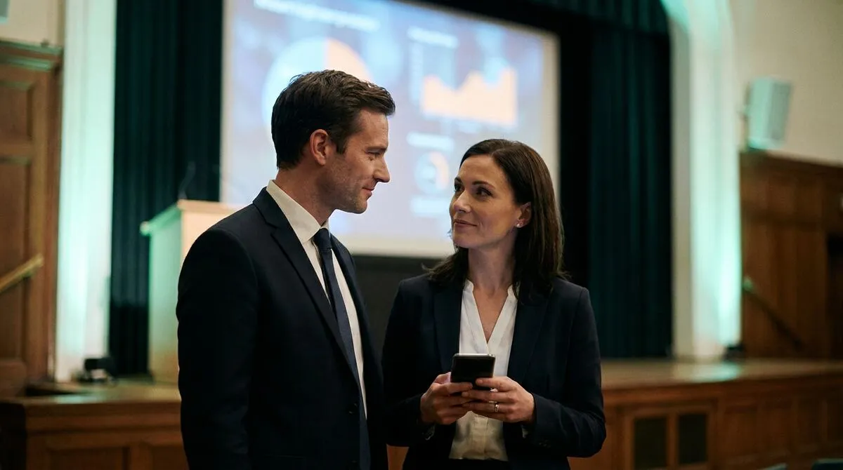 Two presenters exchanging a quiet glance on stage while one checks a device during a brief technical pause