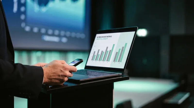 Laptop running a PowerPoint slideshow on a lectern with a presenter checking a phone nearby