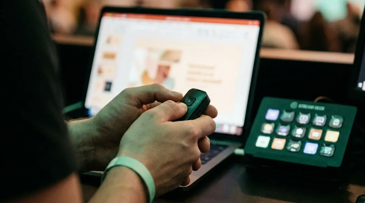 An AV technician tests a phone against a laptop showing a slide deck, a Stream Deck and mixer visible on the desk