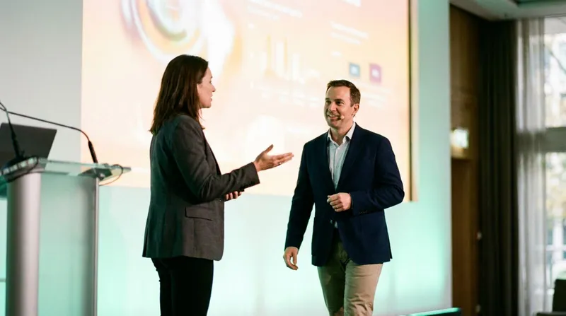 Two co-presenters on stage mid-handoff, one stepping back as the other steps forward to speak