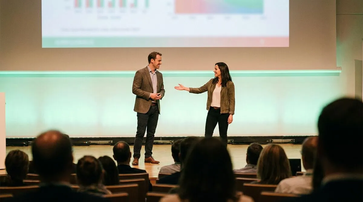 Two co-presenters standing together on stage, one gesturing toward the other during a handoff moment
