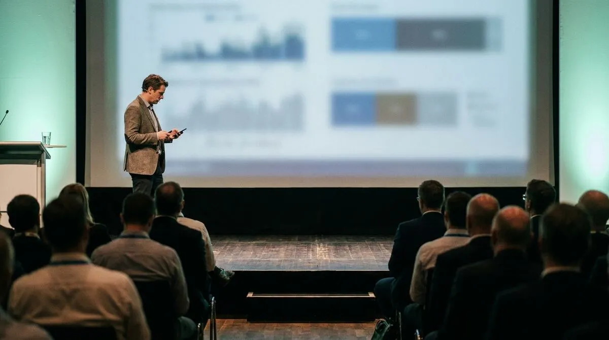 Presenter on a stage glancing at a phone while a large screen behind them shows a softly blurred slide, audience seated in the foreground
