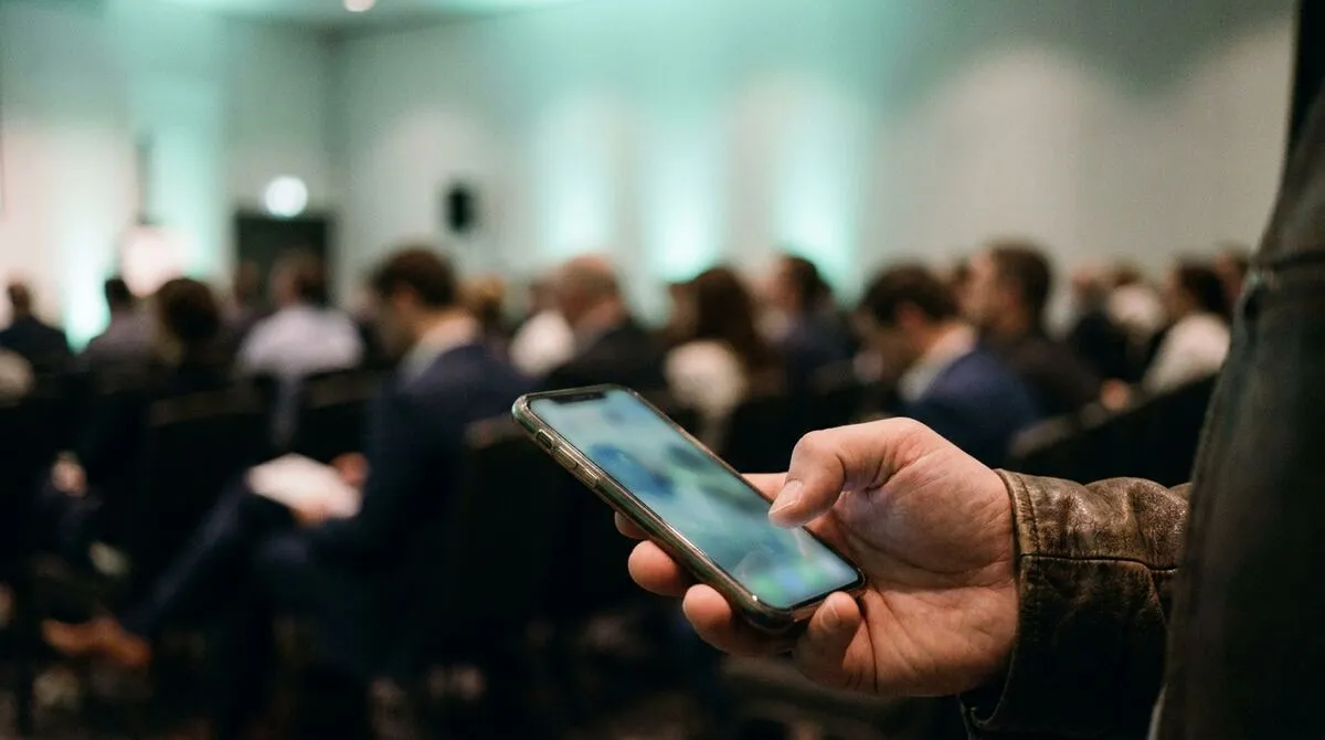 Close view of a speaker's hand holding a phone at waist height with an audience visible softly out of focus behind