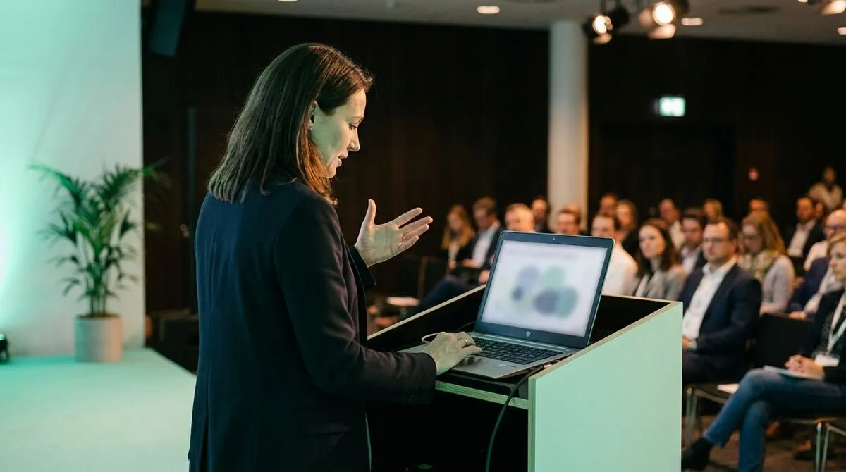 Presenter continuing to speak confidently to the audience while glancing briefly at a laptop showing a paused presentation