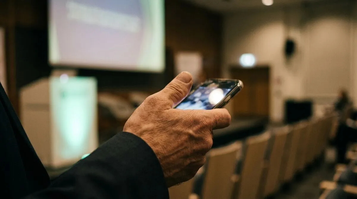 Lecturer standing at the back of a tiered lecture hall holding a phone, slides projected on a large screen at the front
