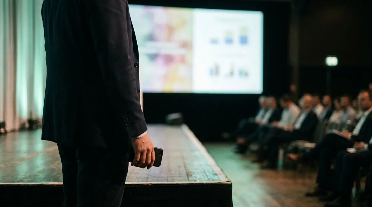 Speaker standing at the edge of a stage close to the front row, phone in hand, away from any lectern or podium
